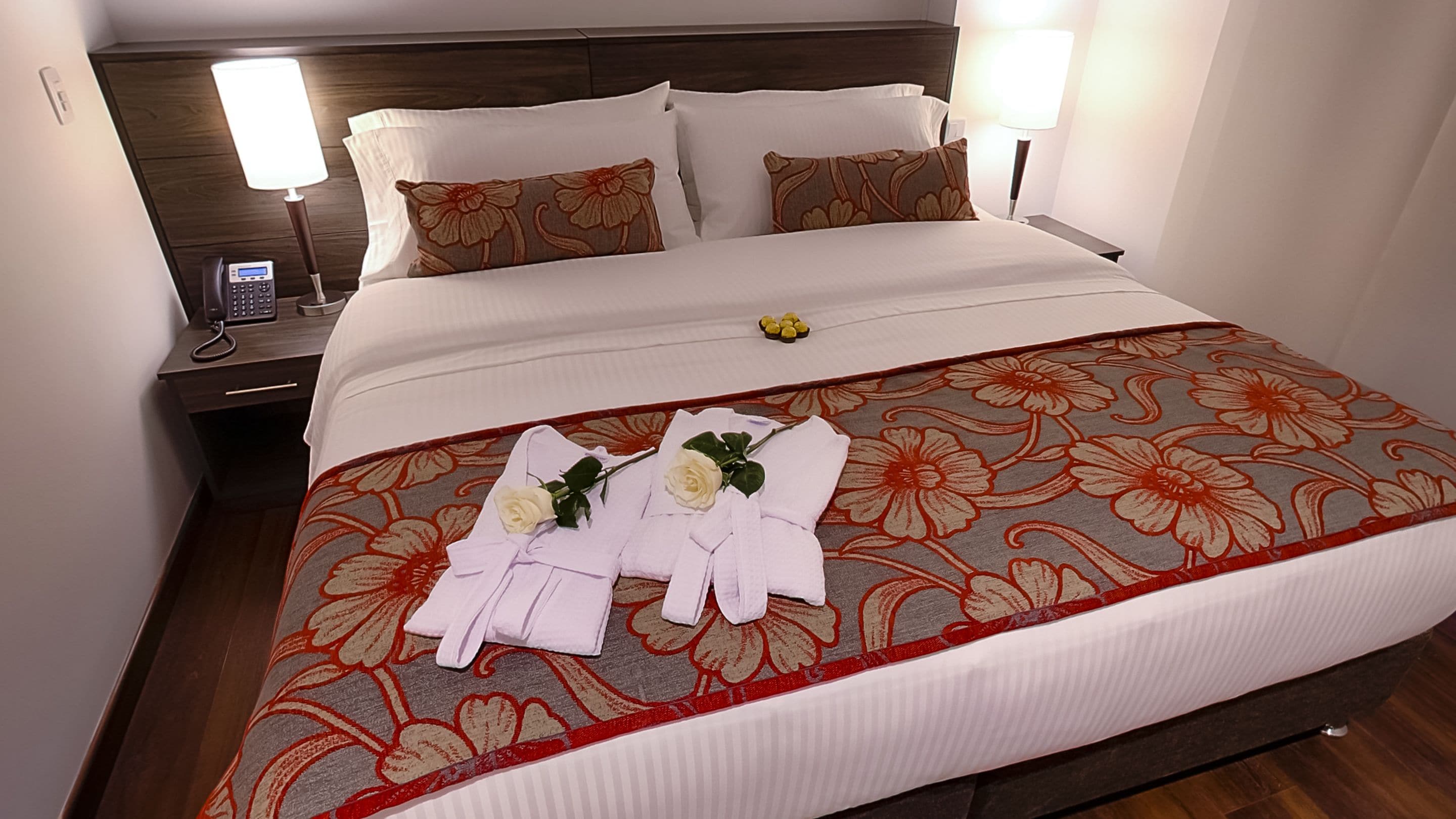 Una cama bien hecha con una colcha floral, dos almohadas y una mesita de noche con un teléfono y una lámpara.