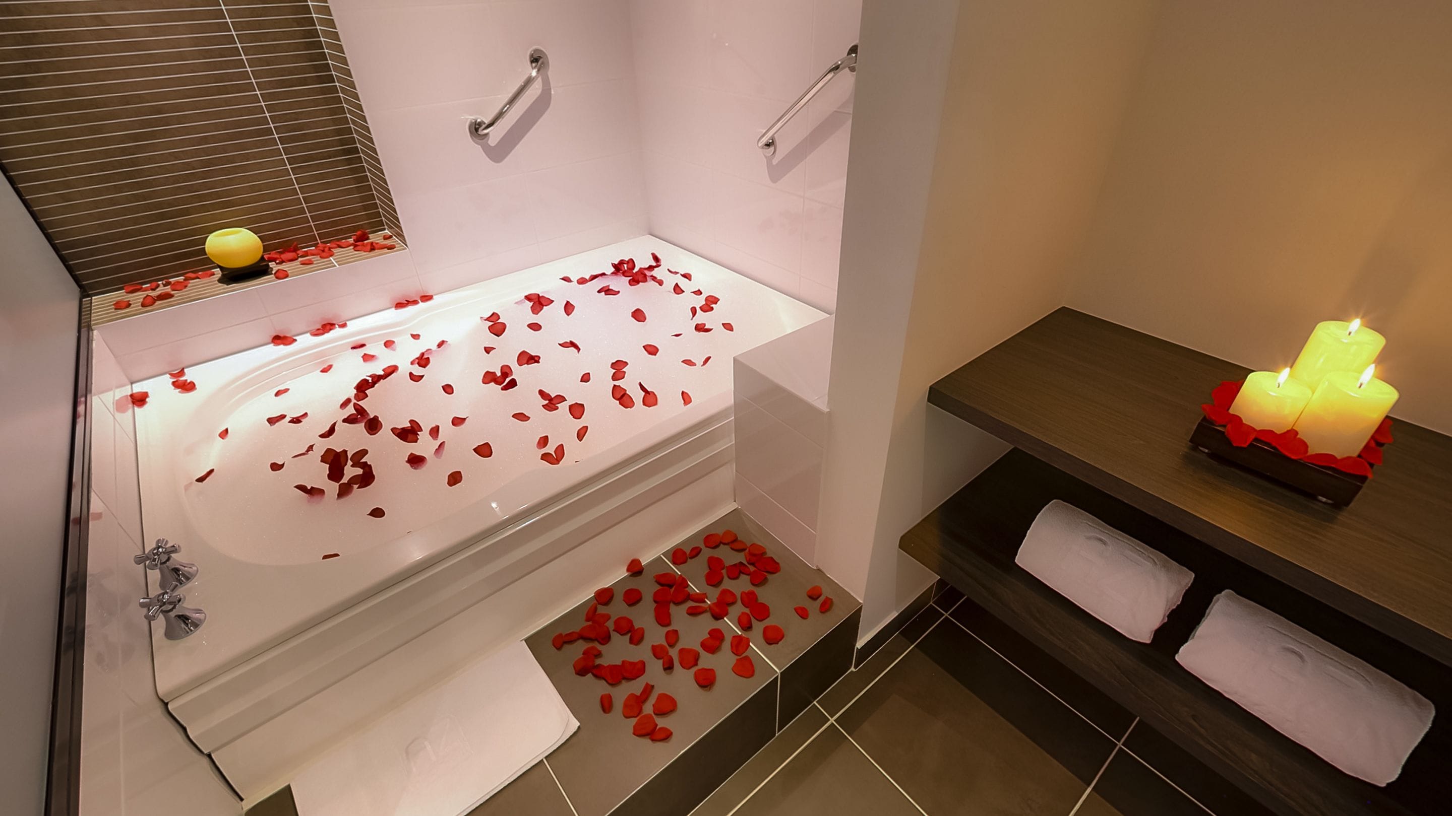 Un baño con una bañera llena de pétalos de rosa, un tocador con una vela y un perchero.