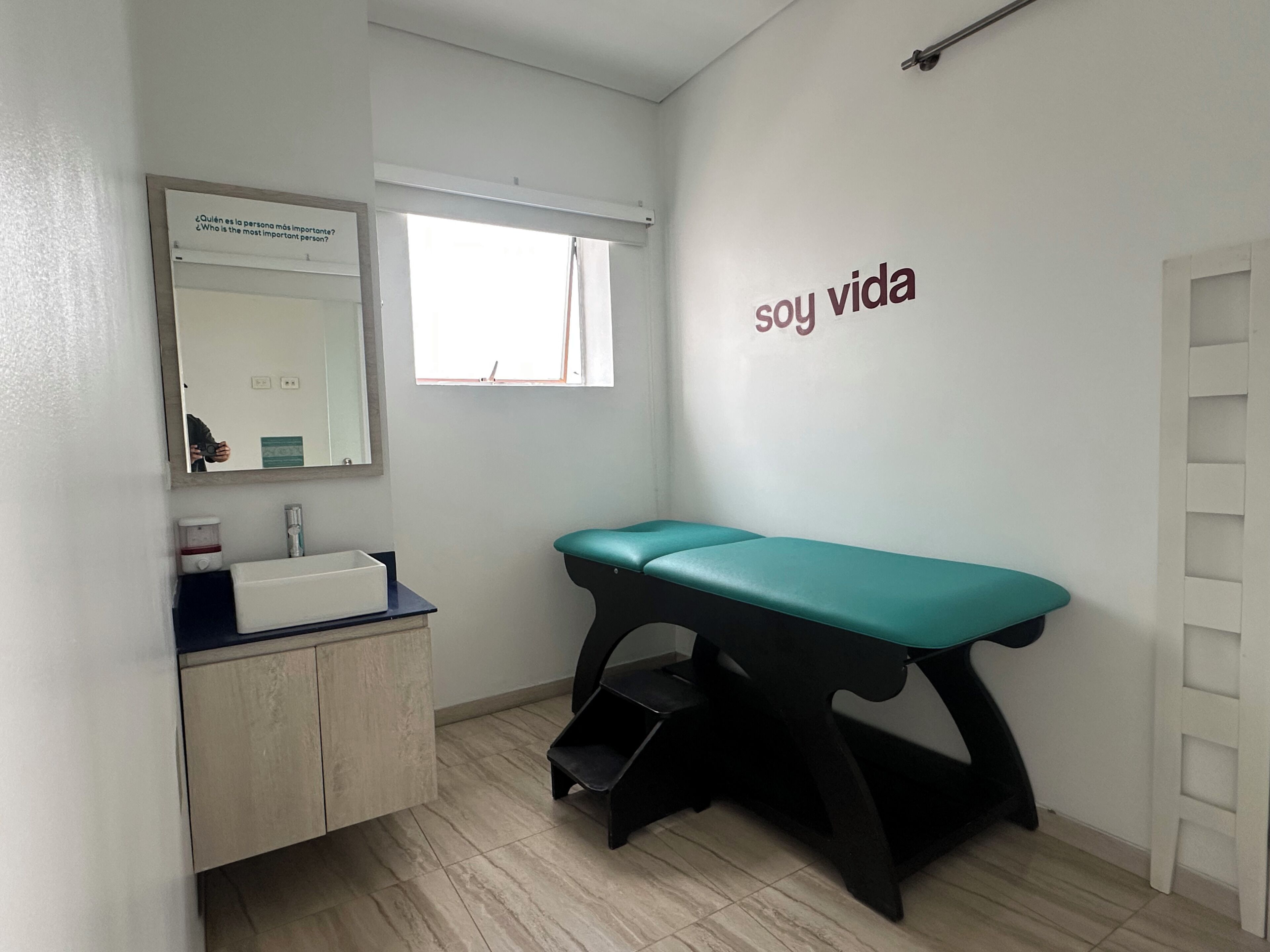 A clinical examination room with a medical examination table, a sink, and a mirror.
