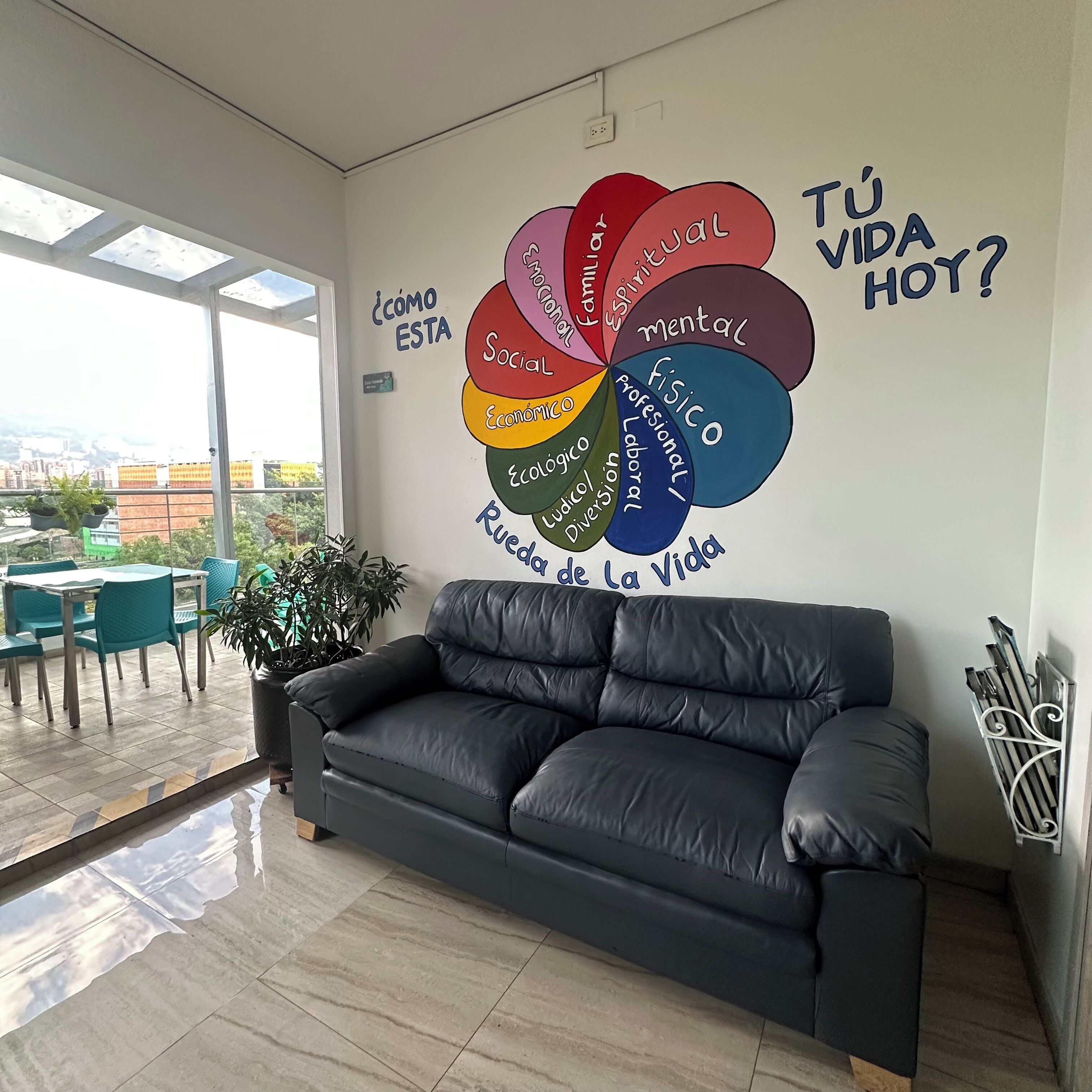 Una sala de estar moderna con un ventanal grande, un sofá de cuero negro y un mural que representa diversos aspectos de la vida.