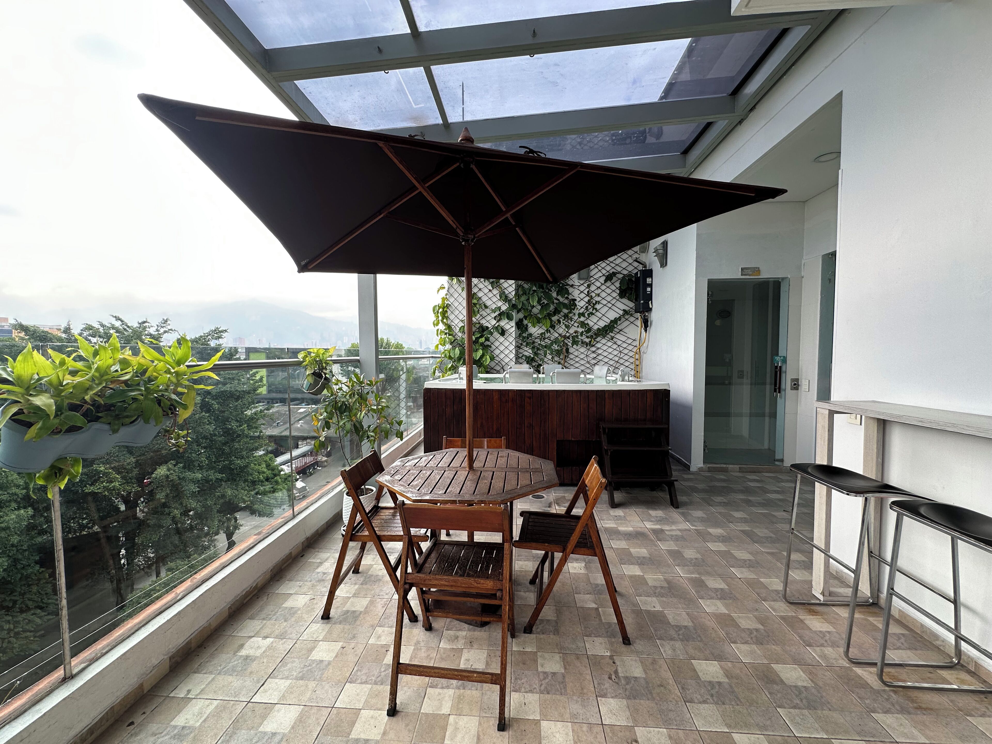 A balcony with a wooden table and chairs, a large umbrella, and a view of the city.