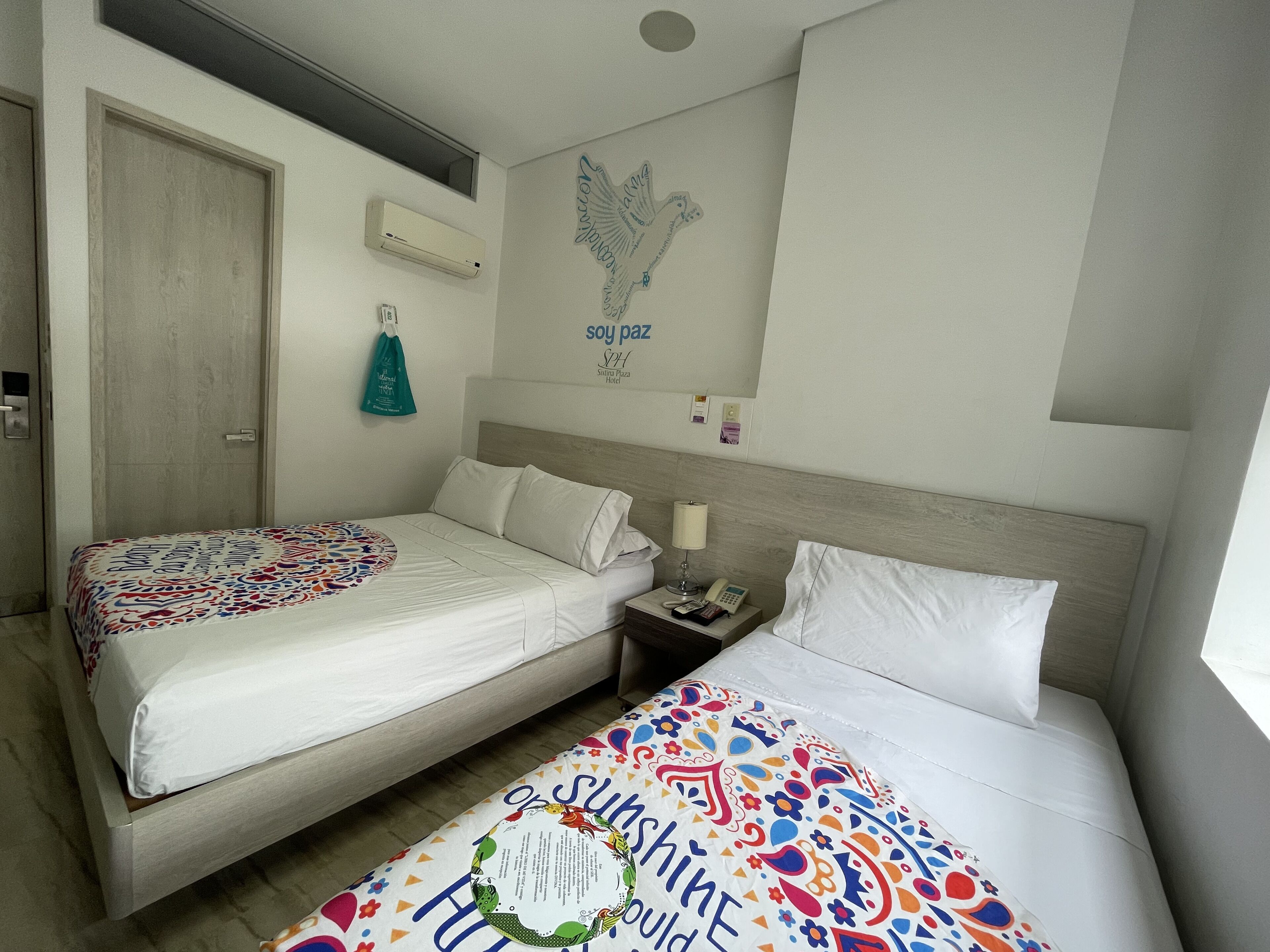 A hotel room with two beds, a wall-mounted air conditioner, a wall clock, and a small table with a lamp and phone.
