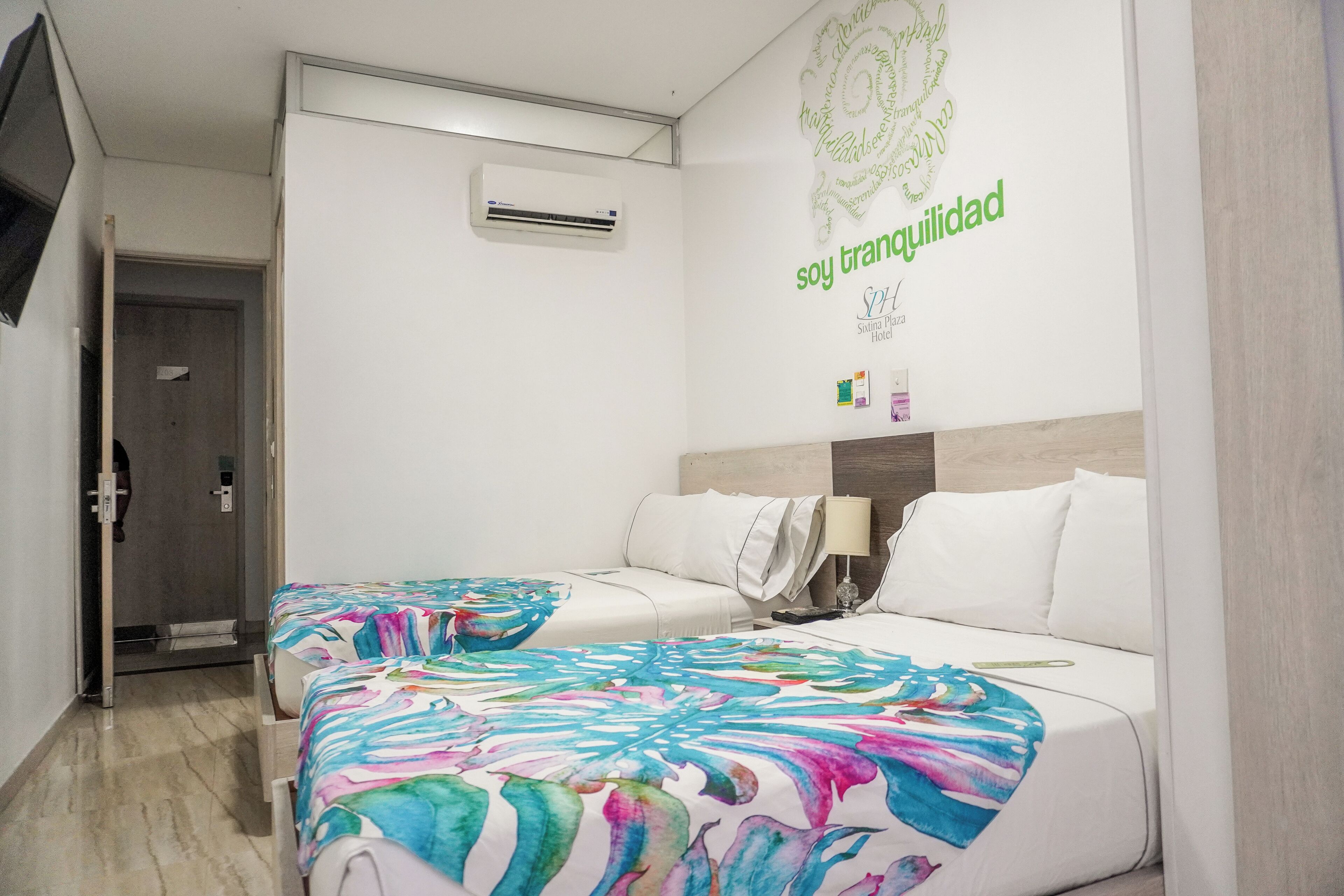 Una habitación de hotel con dos camas, aire acondicionado de pared y una pared decorativa con texto.