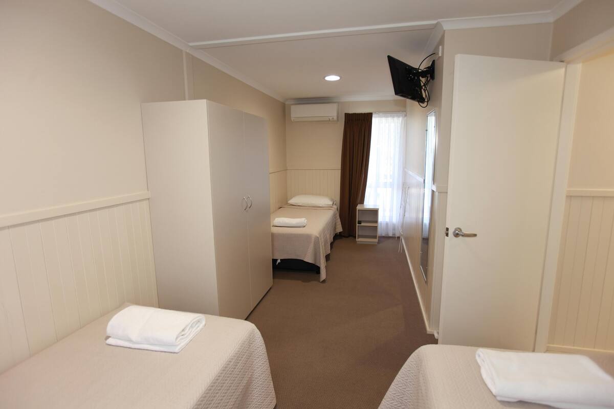 Habitación de hotel con dos camas individuales, un armario, un televisor en la pared y una puerta que da a otra habitación.