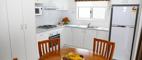 Una cocina moderna con armarios blancos, estufa a gas, microondas y refrigerador. Hay una mesa de comedor con una fuente de frutas en el centro.