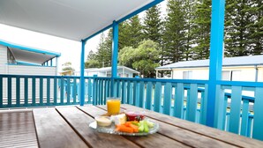 Une table en bois sur laquelle sont disposés un plateau de fruits et de légumes, un verre de jus d’orange et un verre d’eau, le tout sur une terrasse couverte dont les rampes sont bleues.