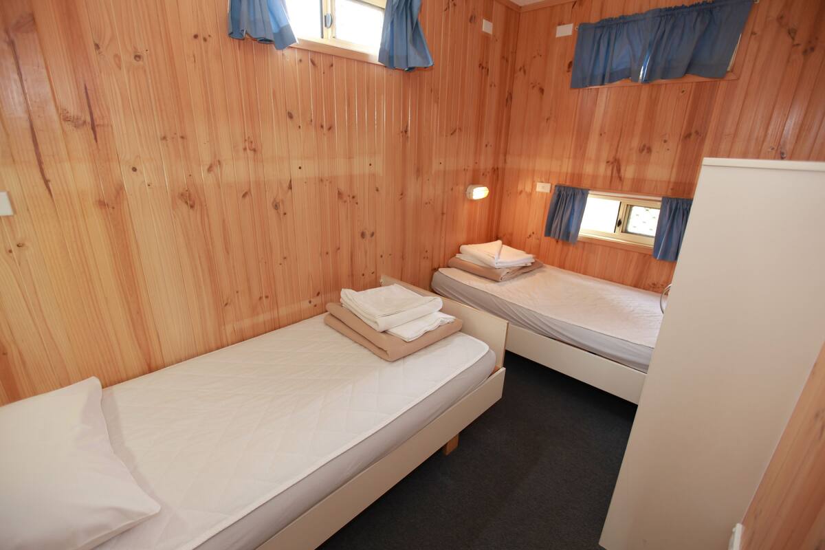 Una habitación pequeña con paneles de madera, dos camas individuales, una mesita y un refrigerador.