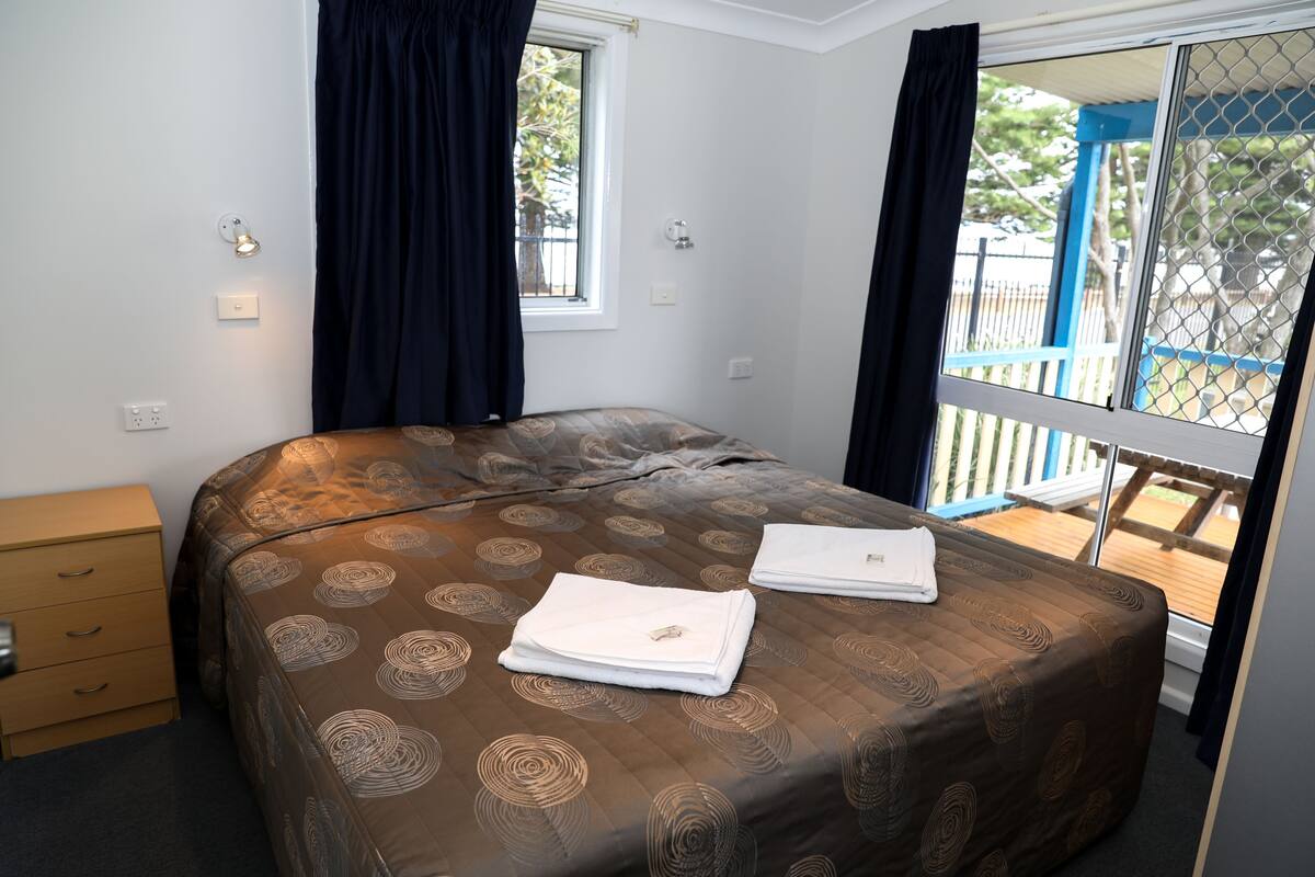 Un dormitorio con cama, mesita de noche, ventana con cortinas y vista a una terraza exterior.