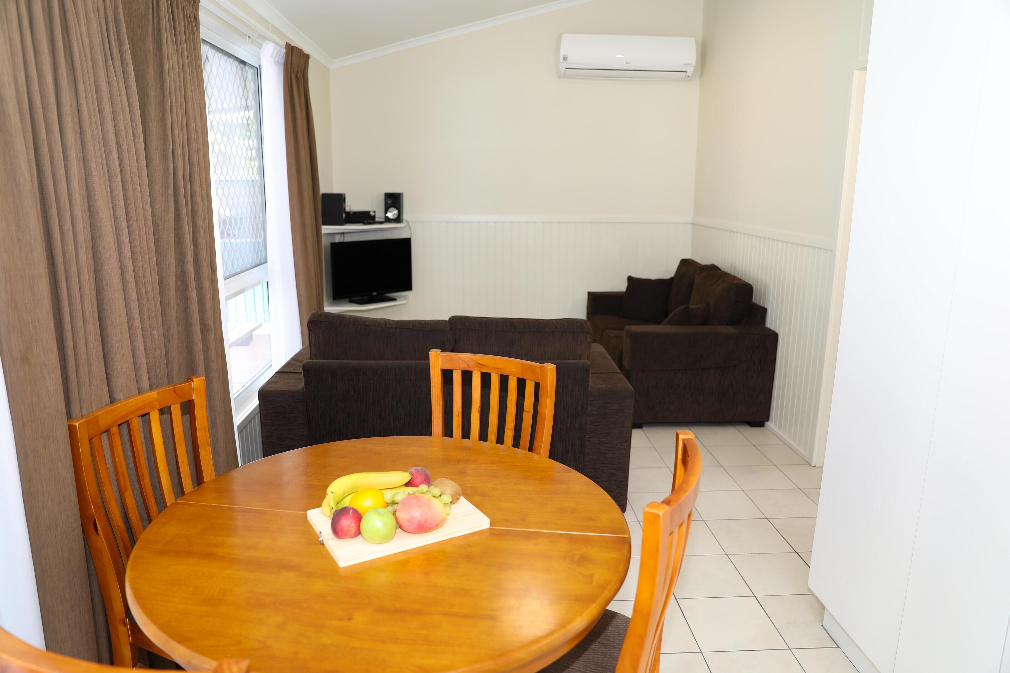 A dining area with a round wooden table, chairs, and a fruit platter. There is a television on a stand and a couch in the background.
