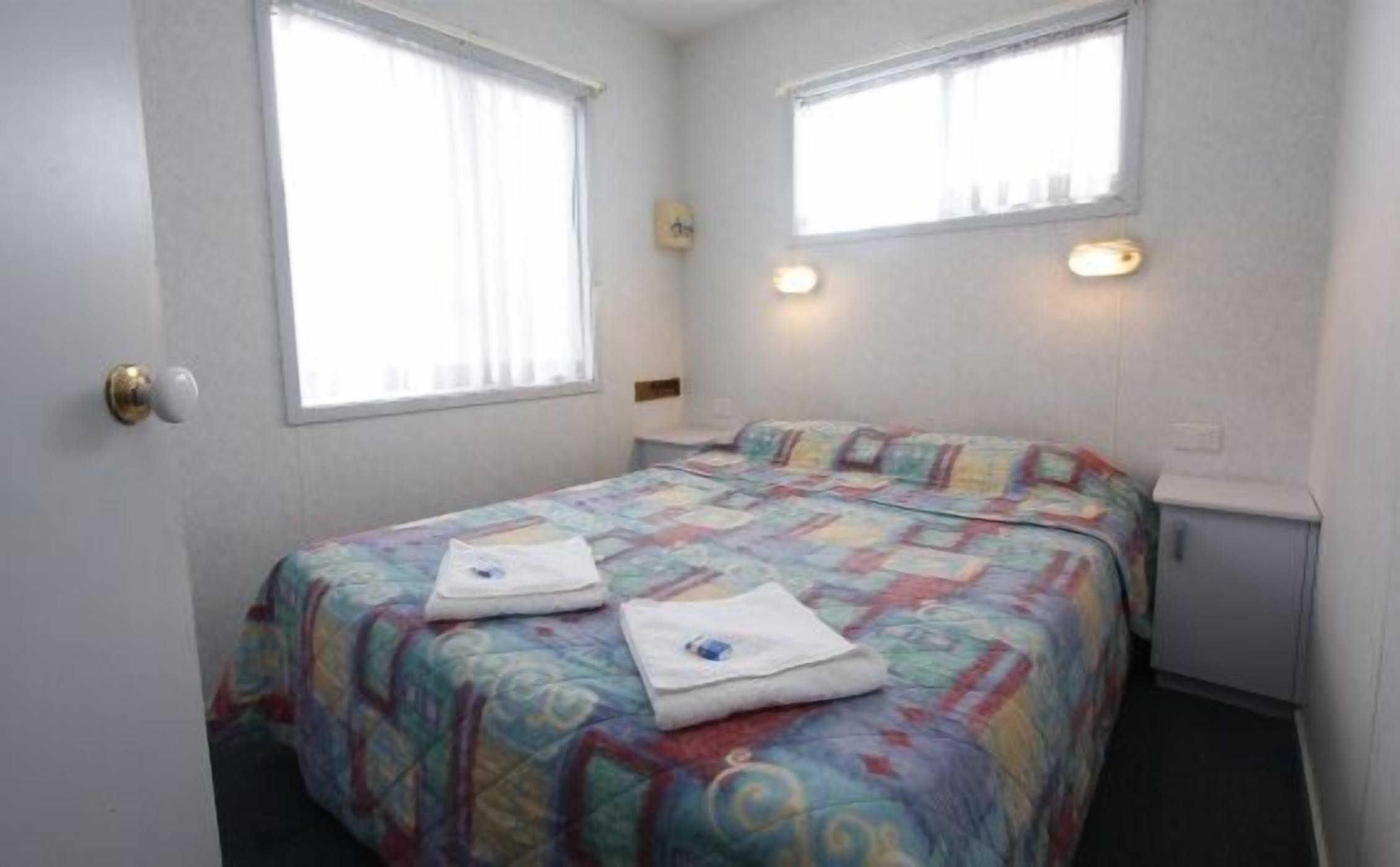 Una habitación pequeña con cama individual, una colcha colorida, dos ventanas con cortinas, una mesita de noche y un armario pequeño.