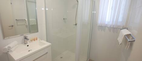 Un baño moderno con ducha envuelta en vidrio, un lavabo blanco con artículos de aseo y un espejo grande.