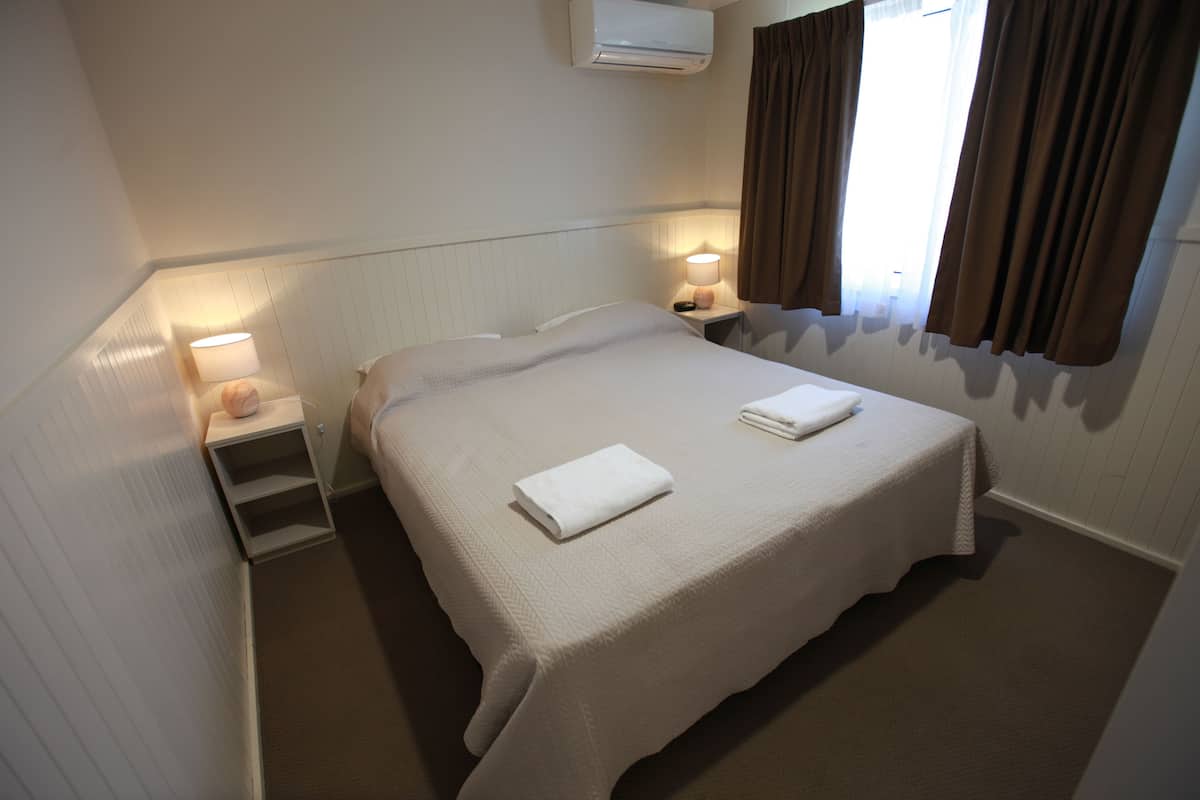 A neatly made bed with a white bedspread and two folded white towels, a bedside lamp, a small shelf with a decorative item, and a window with brown curtains.