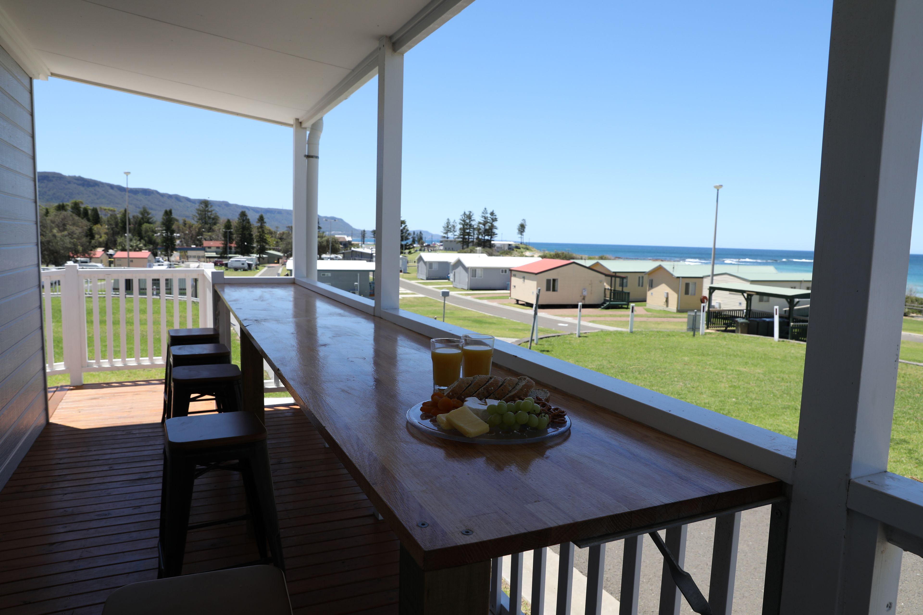 Una terraza de madera con una mesa lista para el desayuno, con vistas a la playa y una zona residencial.