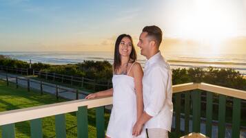 A couple standing on a balcony overlooking a beach with a wooden railing and a grassy area in the foreground.