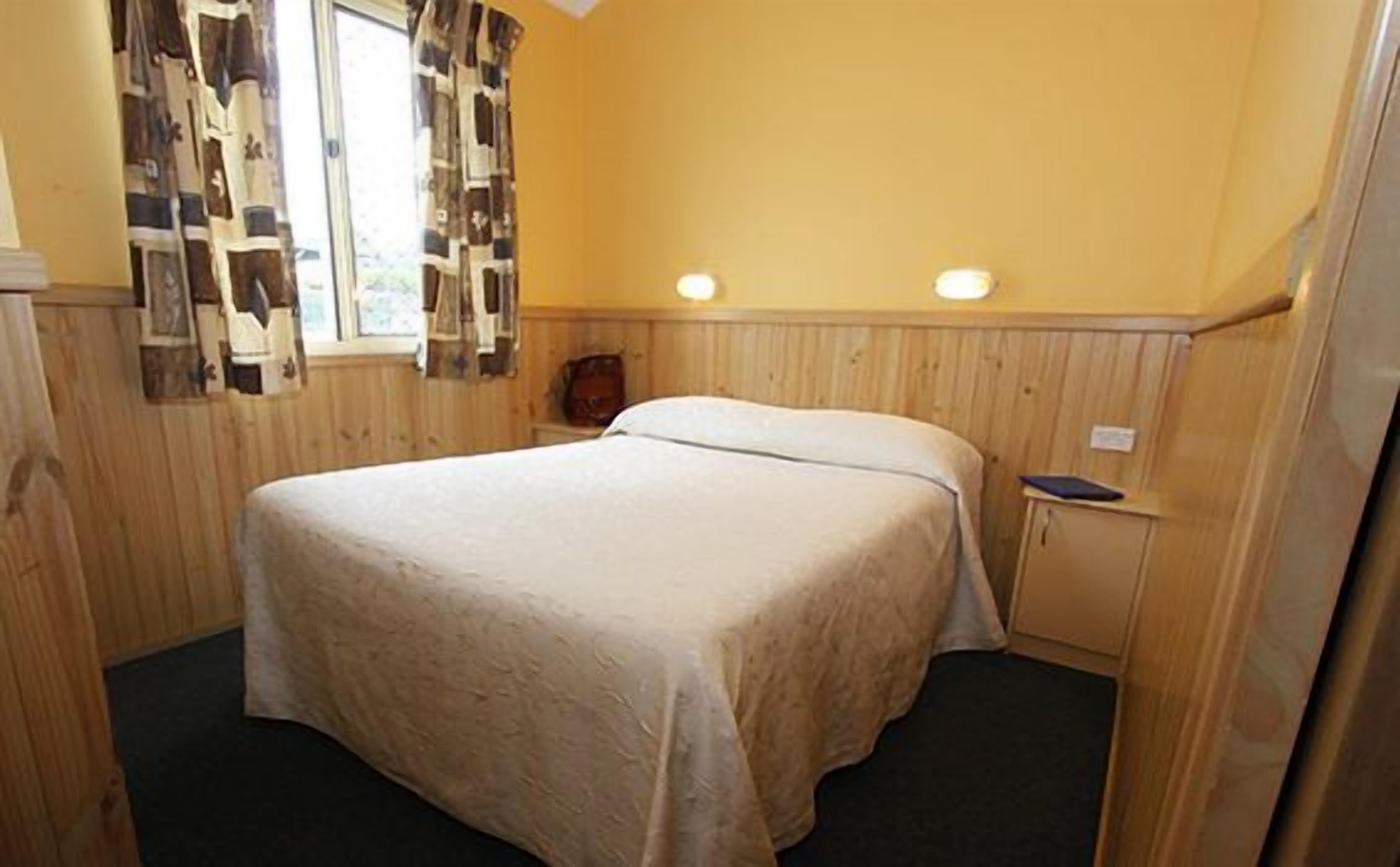 Una cama individual en una habitación con paneles de madera, una mesita de noche pequeña y una ventana con cortinas estampadas.
