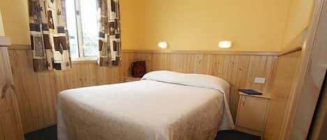 Una cama individual en una habitación con paneles de madera, una mesita de noche pequeña y una ventana con cortinas estampadas.