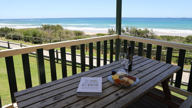 Una taula de fusta amb un llibre, un plat de formatge i una ampolla a un balcó amb vista a una platja.