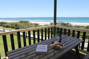 Una mesa de madera con un libro, un plato de queso y una botella en un balcón con vista a la playa.