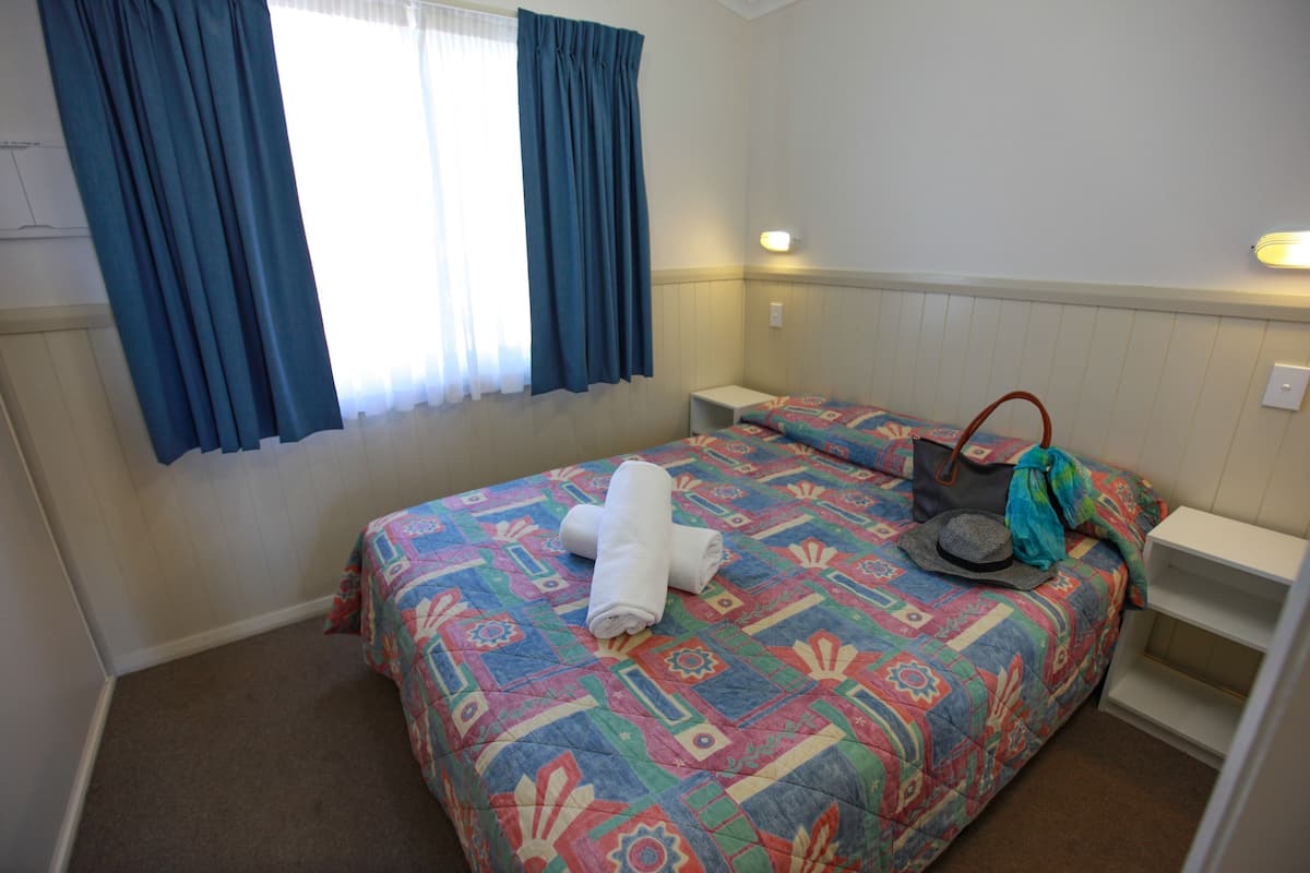 A bedroom with a bed, a nightstand, a bedside table, a window with curtains, and a wall-mounted light.