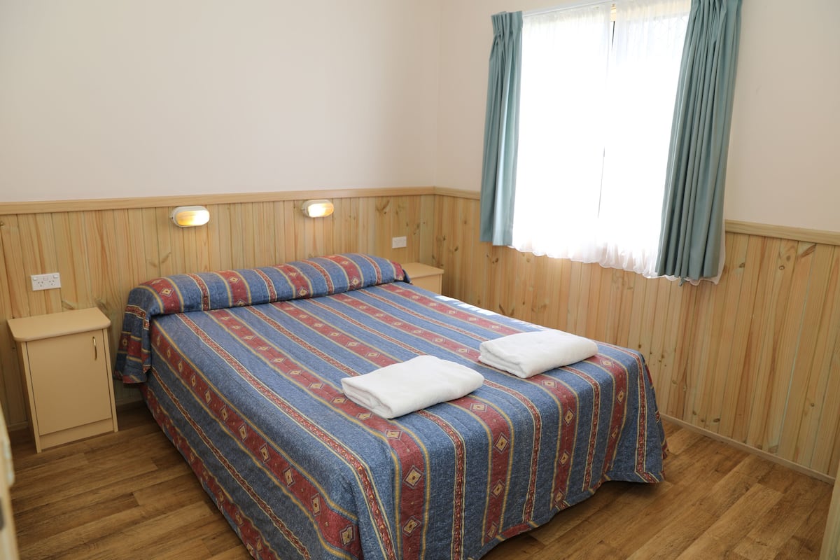A bedroom with a wooden paneled wall, a bed with a striped bedspread, two bedside tables, and a window with curtains.