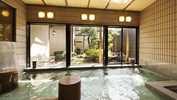 Un onsen japonais traditionnel, avec un bain en pierre, des bancs en bois et une vue sur un jardin, accessible par des portes coulissantes.