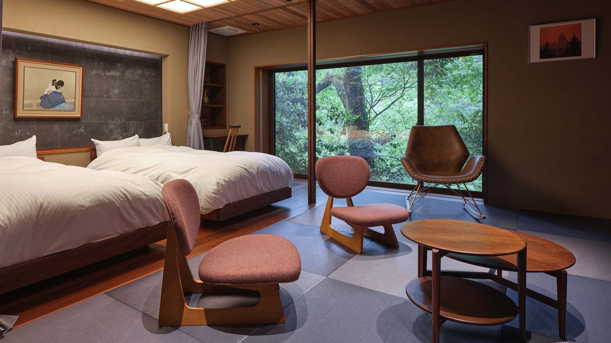 A modern hotel room with a large bed, two chairs, a small table, and a view of greenery through large windows.