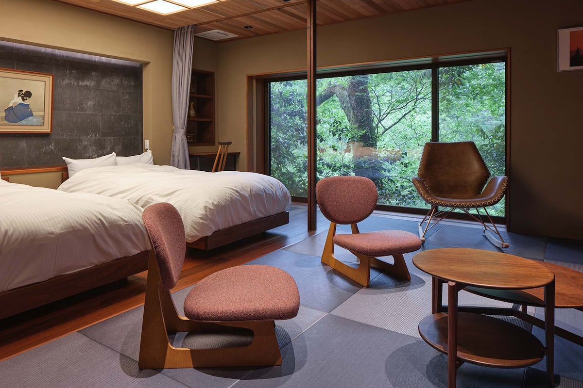 Habitación de hotel moderna con una cama grande, dos sillas, una mesita y vistas a zonas verdes a través de amplios ventanales.