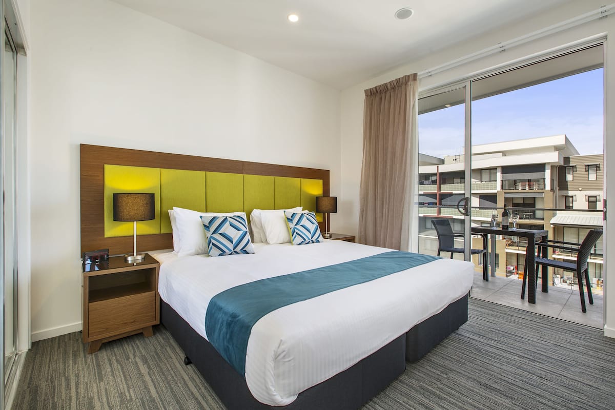 A hotel room with a large bed, a nightstand, a desk with a chair, and a balcony with a view of buildings.