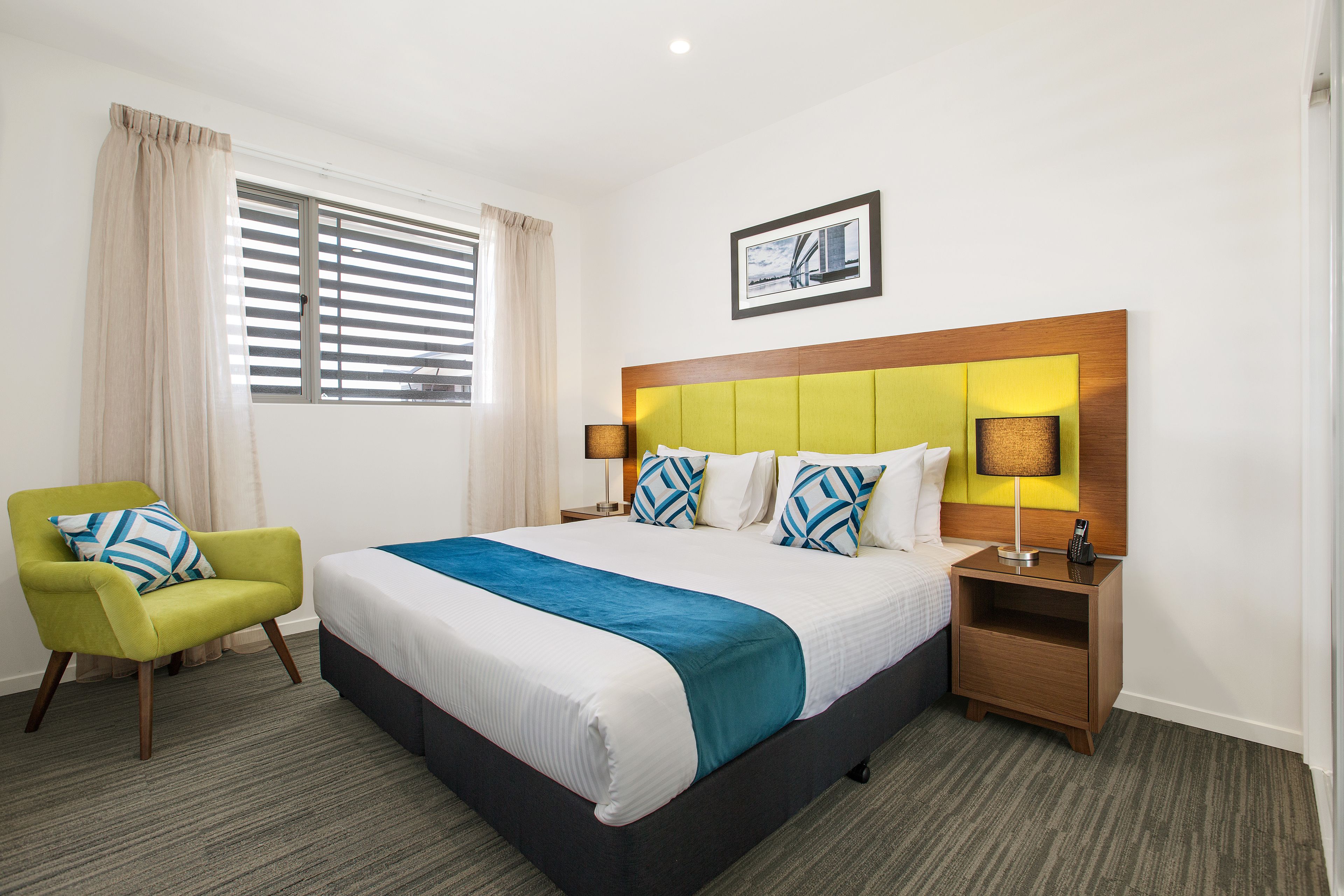 A hotel room with a large bed, a green armchair, a nightstand with a lamp, and a window with blinds.