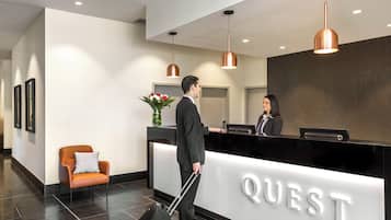 A hotel reception area with a black reception desk, a receptionist, and a guest with a suitcase.