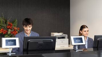 Un mostrador de recepción con dos monitores mostrando el logotipo de "QUEST", una impresora y un jarrón con flores.