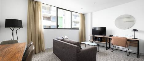 A modern hotel room with a sofa, a desk, a television, and a large window with curtains.