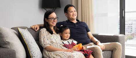 A family sitting on a grey sofa with patterned cushions, a black floor lamp, and a window with a view of buildings.