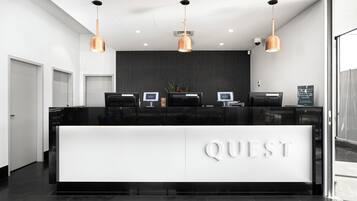 A modern reception desk with the word "QUEST" prominently displayed, three pendant lights, and a dark wall behind the desk.