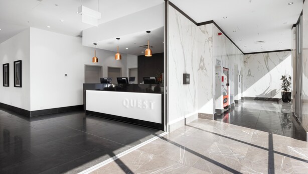 Un área de recepción de hotel moderna con una pared de mármol, un mostrador de recepción identificado con 'QUEST' e iluminación colgante.