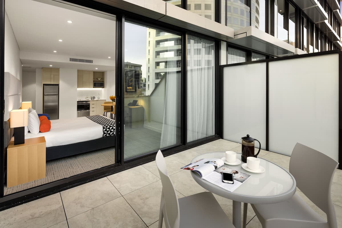 A modern hotel room with a glass wall, a bed, a small kitchenette, and a balcony with a table and chairs.