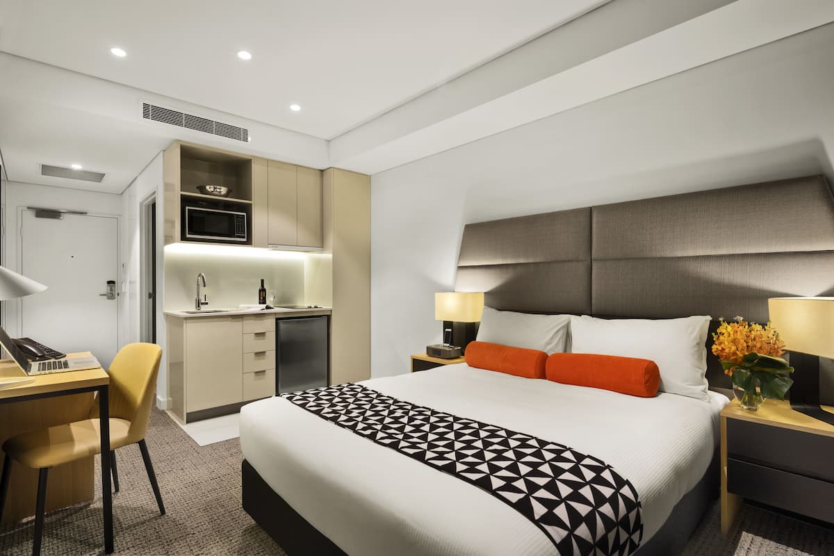 A modern hotel room with a large bed, a desk with a computer, a kitchenette, and a bathroom.
