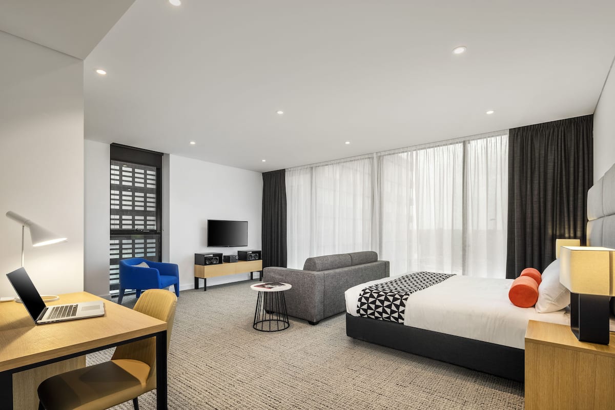 A modern hotel room with a bed, a desk, a chair, a sofa, a TV, and a small table.