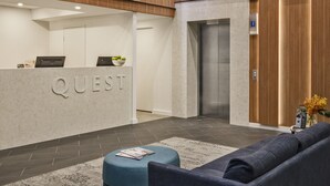 A modern reception area with a 'QUEST' sign, a reception desk, a blue sofa, and an elevator. - Quest St Leonards (St. Leonards)