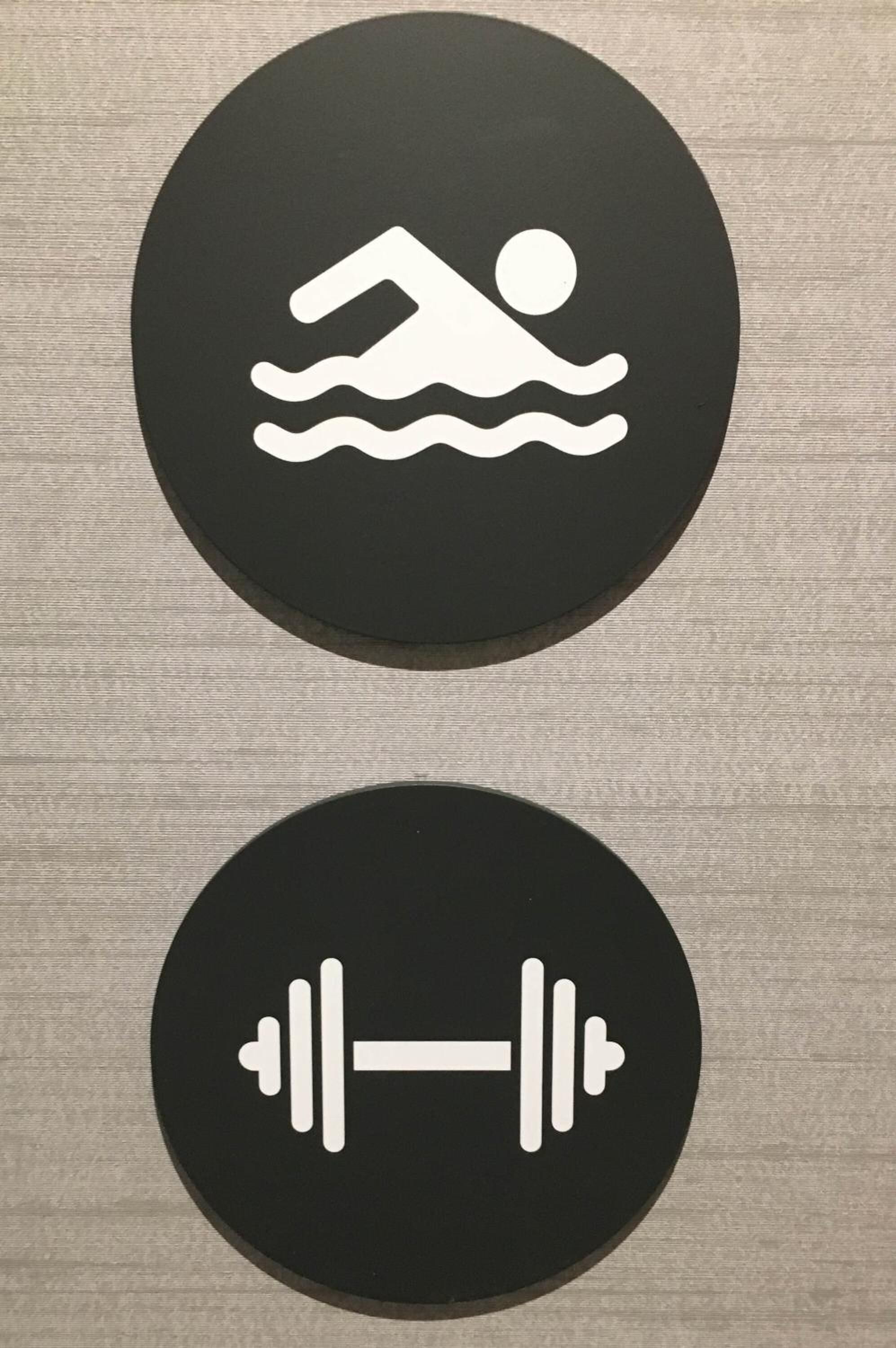 Två runda skyltar, den ena visar en person som simmar och den andra en skivstång, vilket troligen indikerar en pool och ett område för styrketräning.