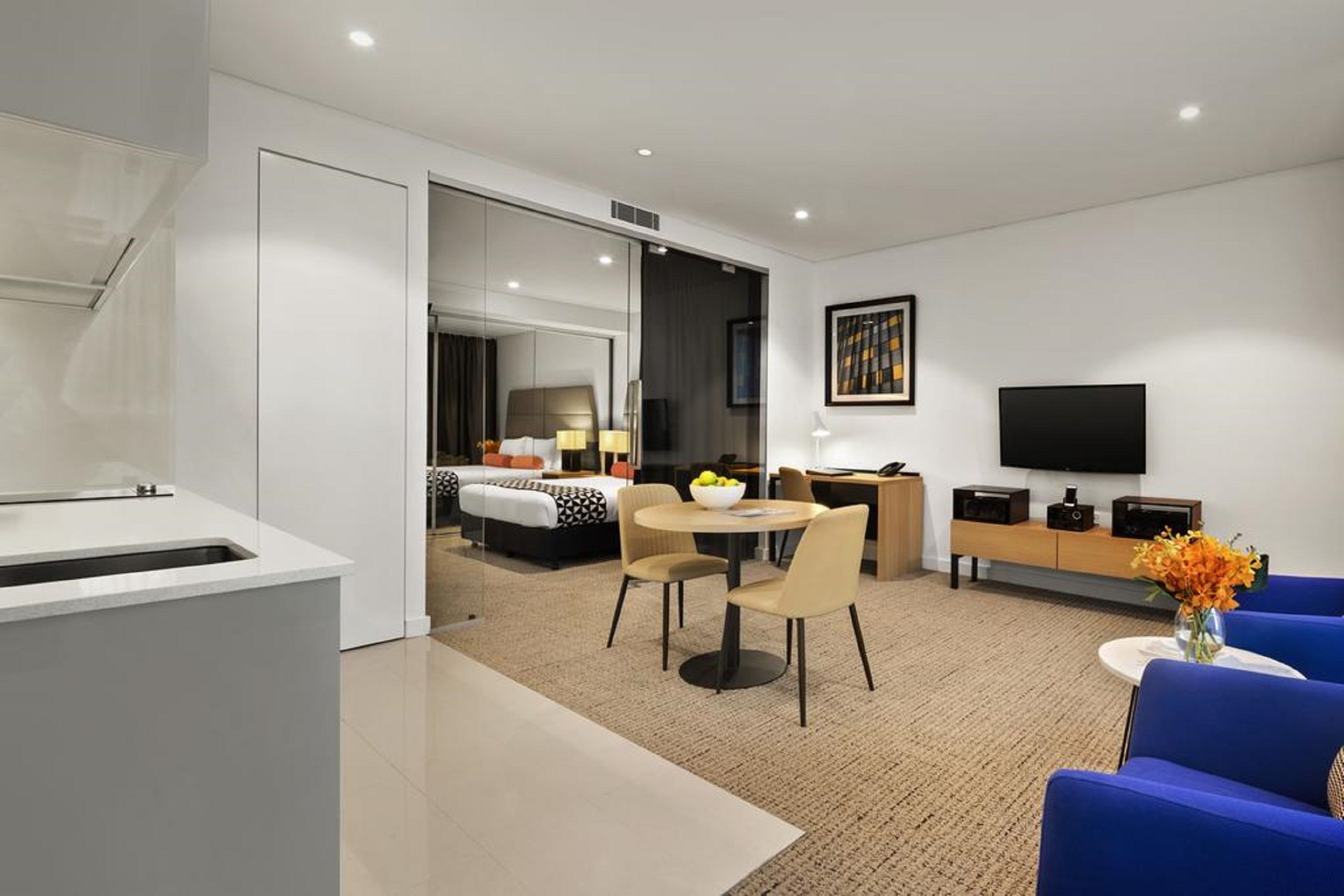 A modern hotel room with a kitchenette, a dining area, a living area with a TV, and a bedroom with a large bed and a desk.