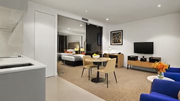 A modern hotel room with a kitchenette, a dining area, a living area with a TV, and a bedroom with a large bed and a desk.