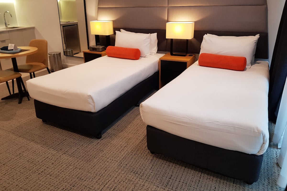 A hotel room with two beds, a desk, a chair, a trash bin, and a lamp.