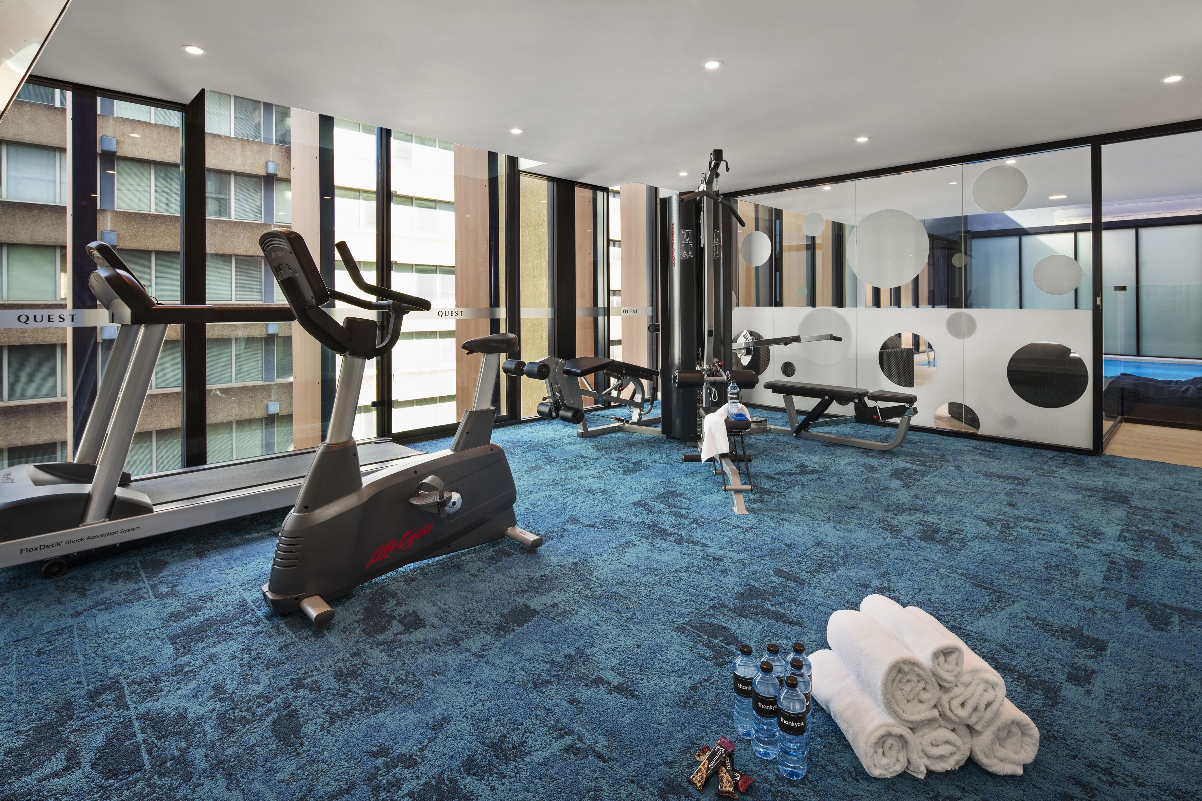 A modern hotel gym with cardio equipment, a weight bench, and a large window offering a view of the city.