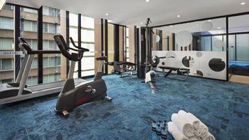 A modern hotel gym with cardio equipment, a weight bench, and a large window offering a view of the city.