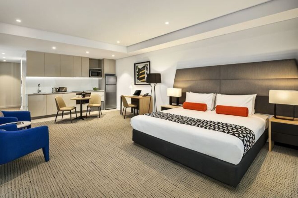 A hotel room with a large bed, a desk, a chair, a sofa, and a kitchenette.