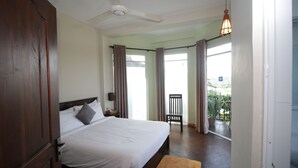 Ein Schlafzimmer mit einem großen Bett, einem Deckenventilator, einem Fenster mit Vorhängen, einem Sessel und Blick ins Freie.