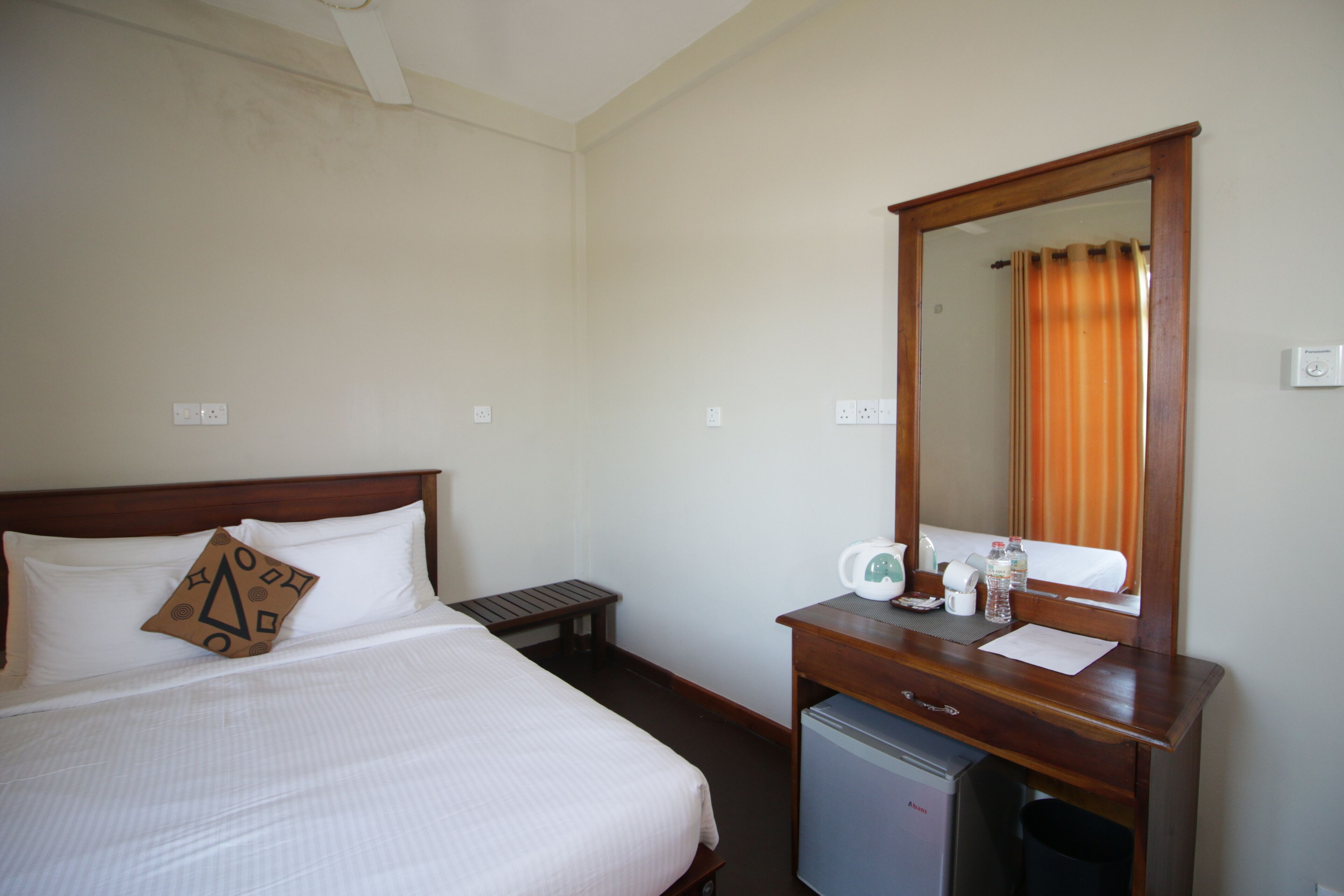 Habitación de hotel con cama, un pequeño escritorio con espejo, refrigerador y ventana con cortinas.