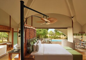 Quarto com uma cama grande, ventilador de teto, vista para uma piscina e área de refeições com cadeiras e mesa.