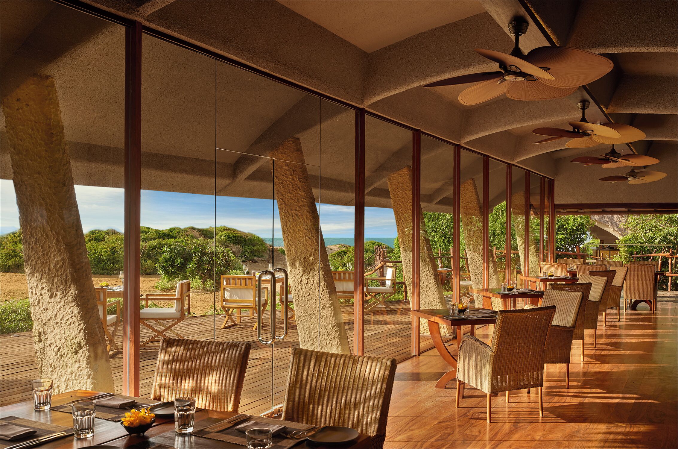 A spacious outdoor dining area with wooden tables and chairs, large windows, and ceiling fans.
